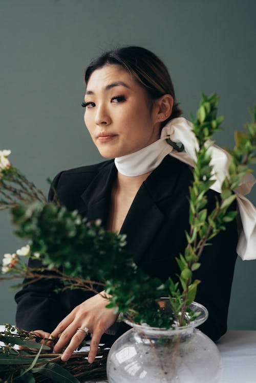 Ethnic female with silky scarf on neck looking at camera at table with fresh green branches on gray background