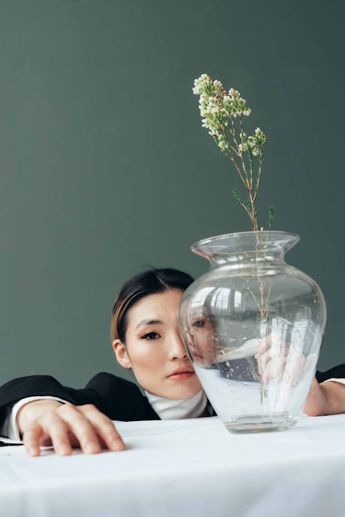 Free Ethnic female with dyed hair looking at camera at white table with glass vase with fragile flowers on gray background Stock Photo