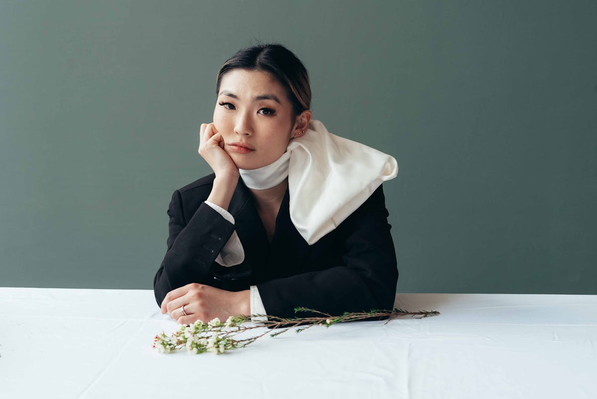 Peaceful bored ethnic female in stylish black formal jacket at table with fragile blooming branch on gray background