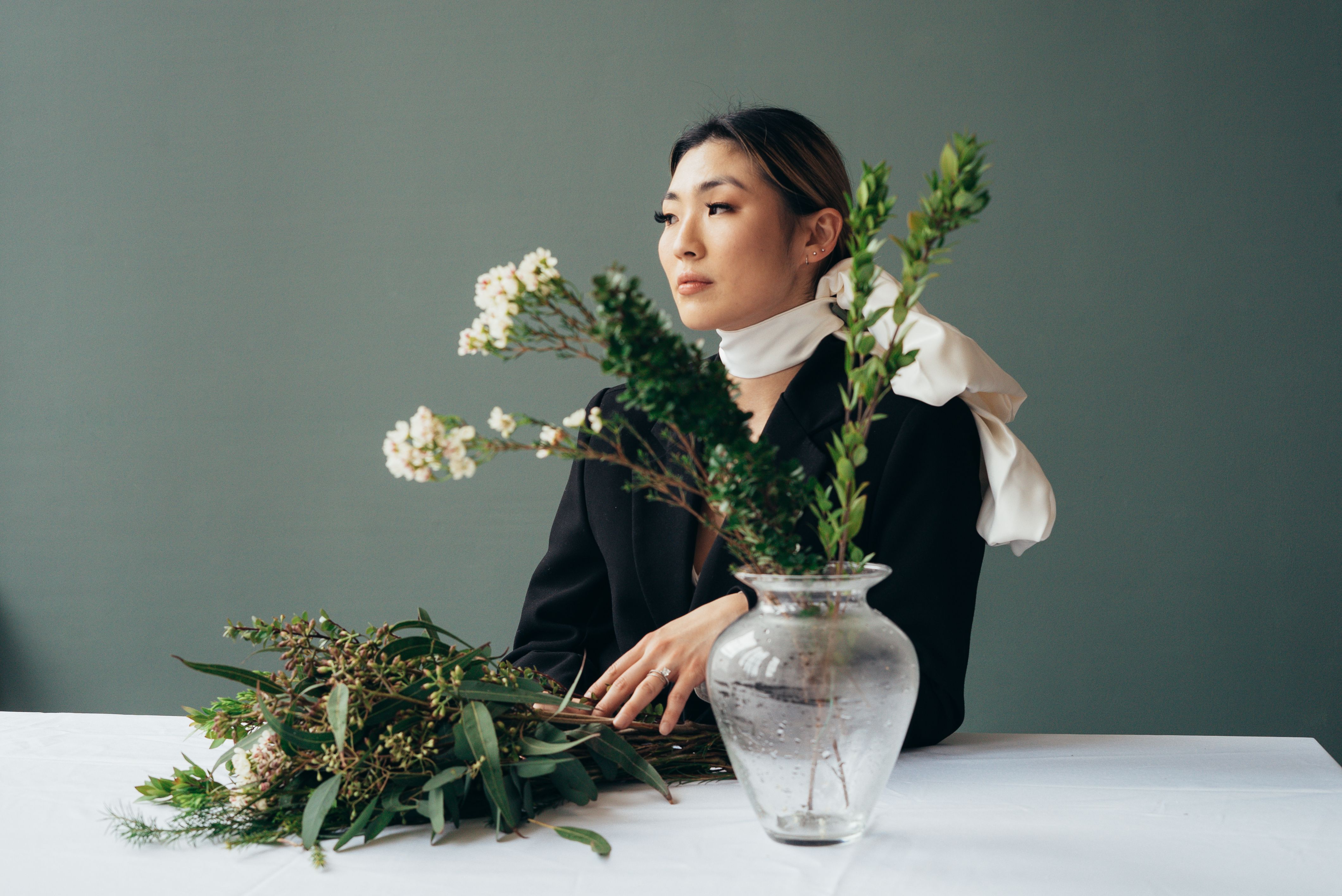 Gentle ethnic female in stylish black jacket looking away at table with blooming white flowers in glass vase with water