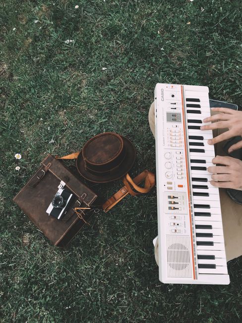 From above of crop unrecognizable male musician playing song on synthesizer while sitting on grassy ground near hat and photo camera