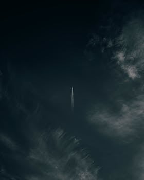 A jet airplane flying high with a visible contrail against a dark, cloudy sky.