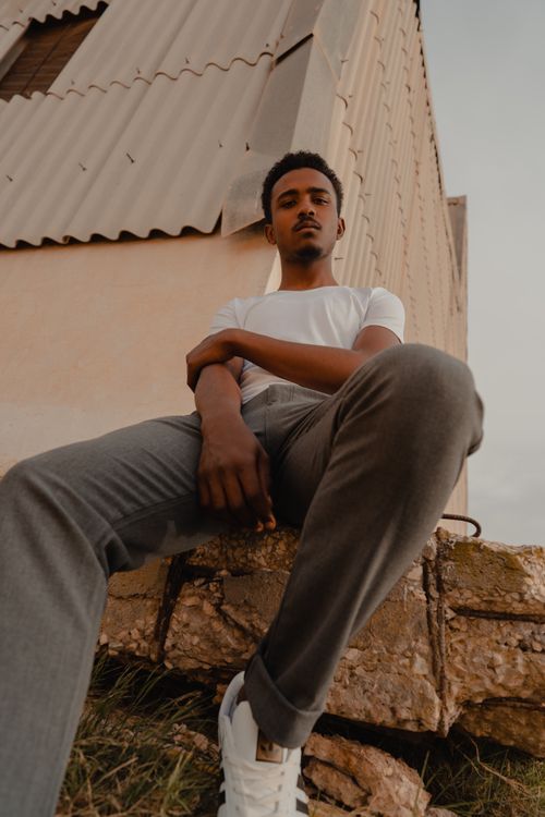 Free Confident young man in a white shirt and jeans sits against a rustic building, portraying modern style. Stock Photo