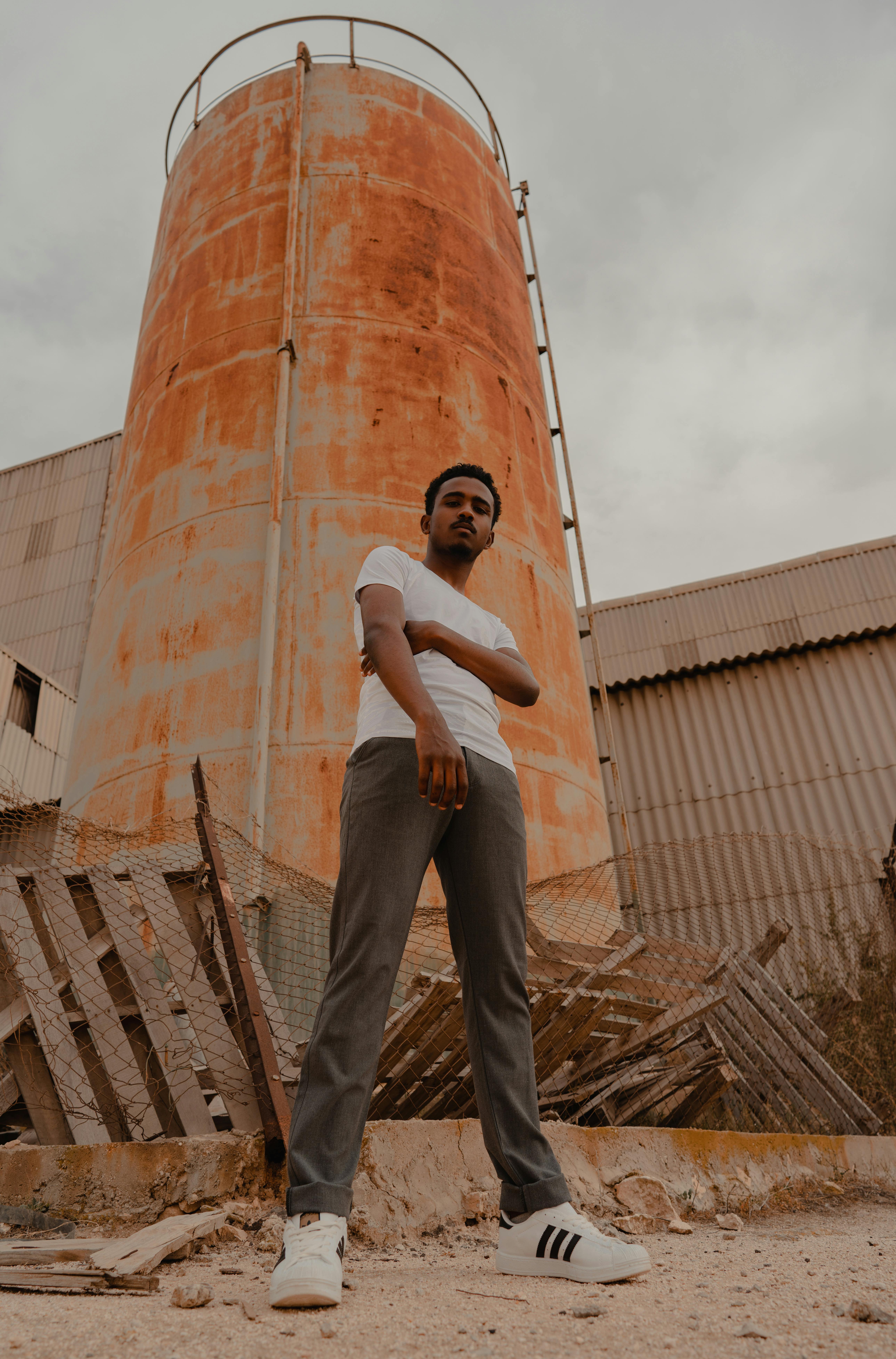 Free Stylish man in a white shirt and gray pants poses confidently by a large industrial smokestack. Stock Photo