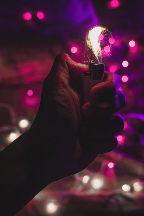 Crop unrecognizable person with burning lighter in hand standing in dark room with glowing lights of garland on blurred background