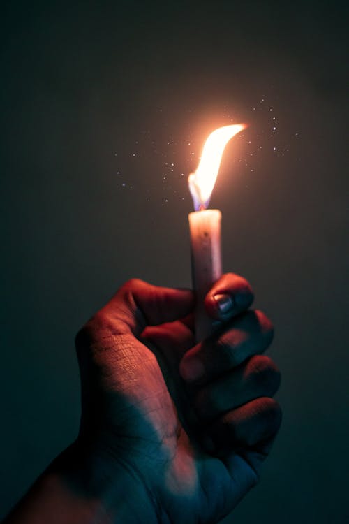 Crop unrecognizable person with small white burning candle with bright sparks in hand standing on dark background in low light