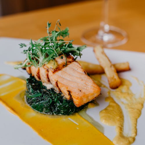 Closeup of delicious grilled salmon fillet and spinach served on white plate with french fries and sauce on table in restaurant