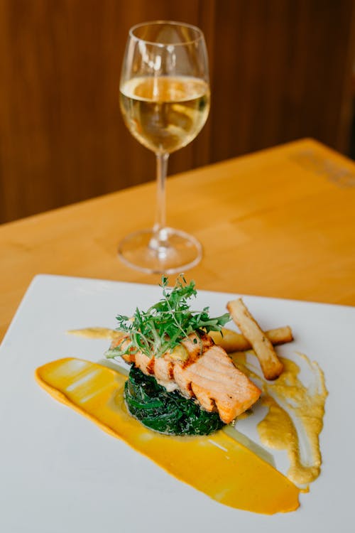From above of delicious appetizing salmon steak and spinach decorated with arugula served on plate with fries and sauce near wineglass in restaurant