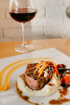 From above of appetizing grilled beefsteak with potato puree and vegetables served on wooden table with glass of red wine in restaurant