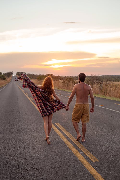 Back view of unrecognizable couple holding hands and walking on asphalt roadway placed among fields in countryside at sunset time