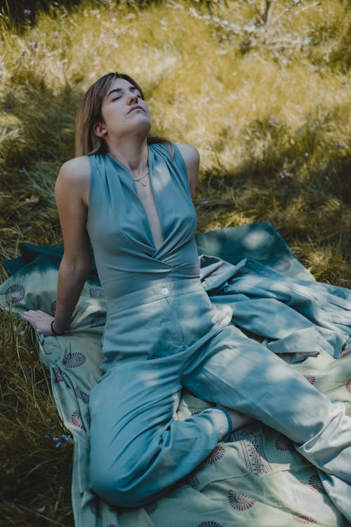 From above of young peaceful female leaning on hands while sitting on blanket spread on grassy field with closed eyes