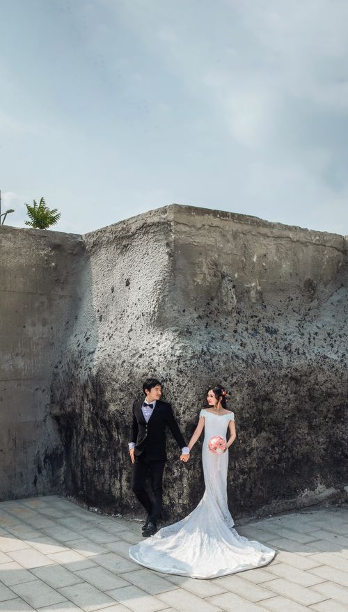 Free Wedding couple in elegant attire posing against a textured concrete wall outdoors. Stock Photo