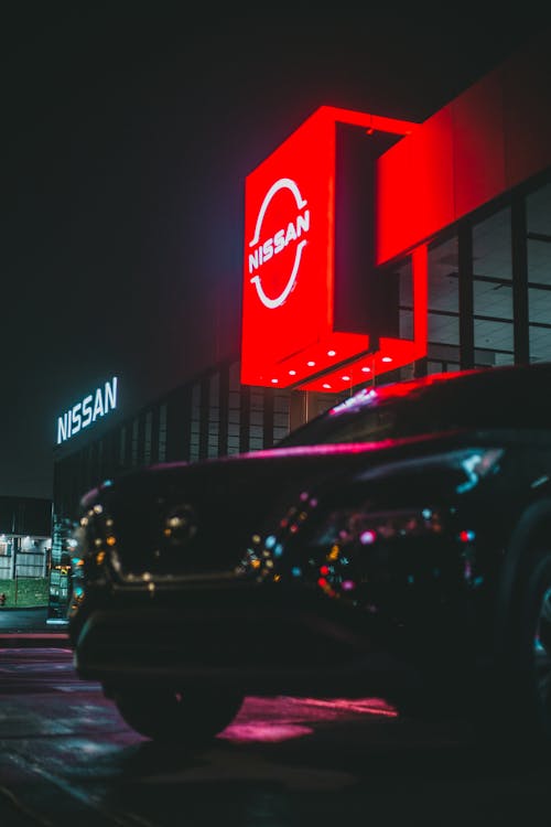 Expensive automobile on parking lot near building of car dealership with glowing red signboard against night sky