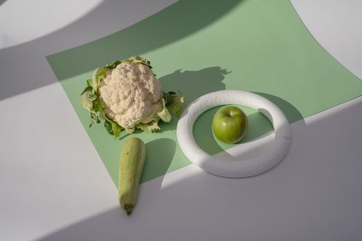 A minimalist still life featuring a green apple, zucchini, and cauliflower on a soft background.