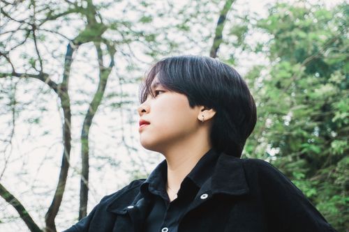 Free Side view portrait of a young woman with short hair, outdoors amidst green trees, looking thoughtfully. Stock Photo