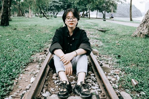 Free Asian woman in glasses sitting on railway in green park, wearing black jacket and jeans. Stock Photo