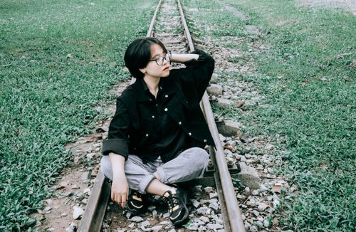 Free A stylish individual in a black jacket sits on railway tracks, surrounded by green grass. Stock Photo