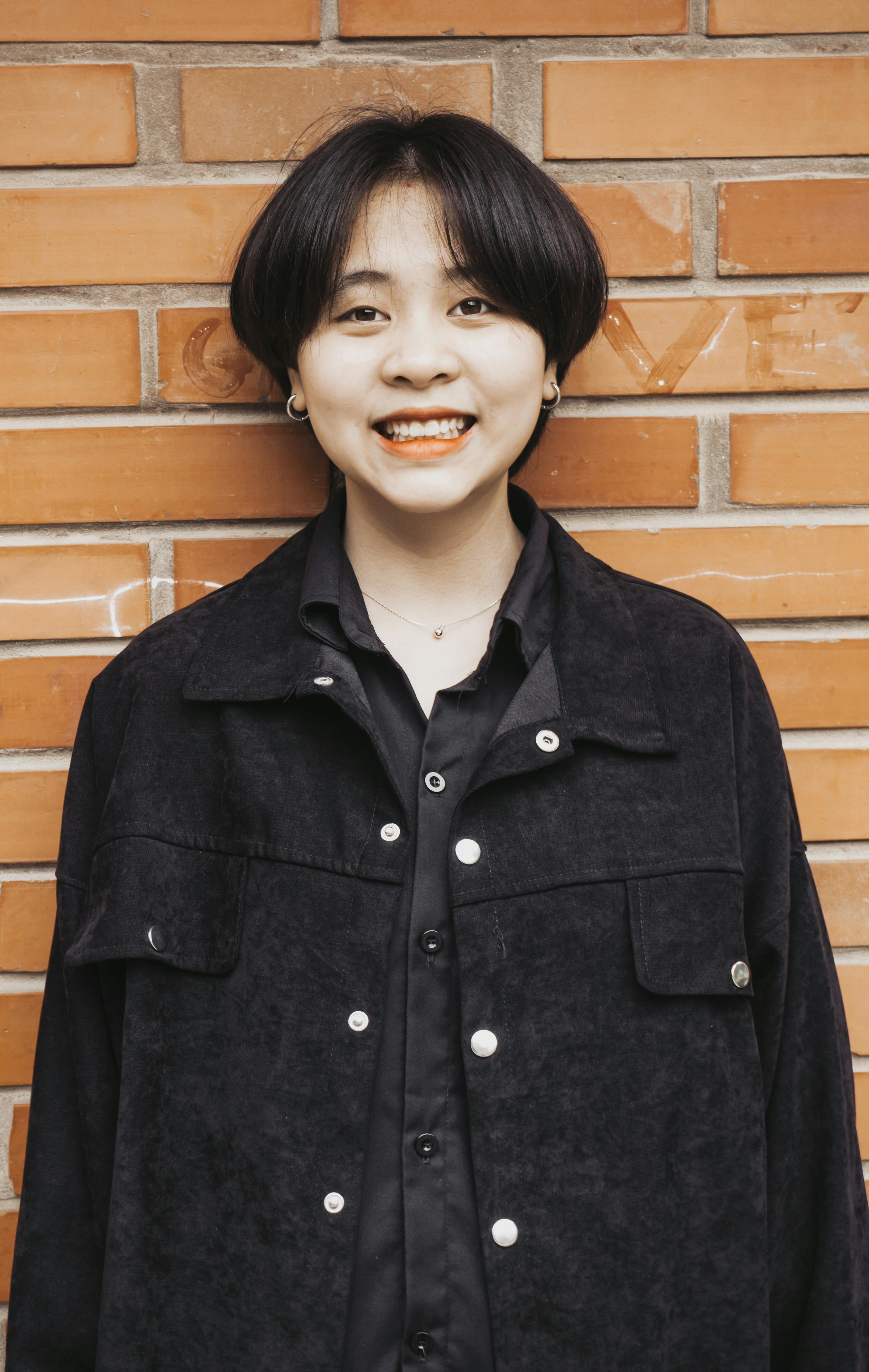 Smiling woman with short hair posing in stylish jacket against a brick wall background.