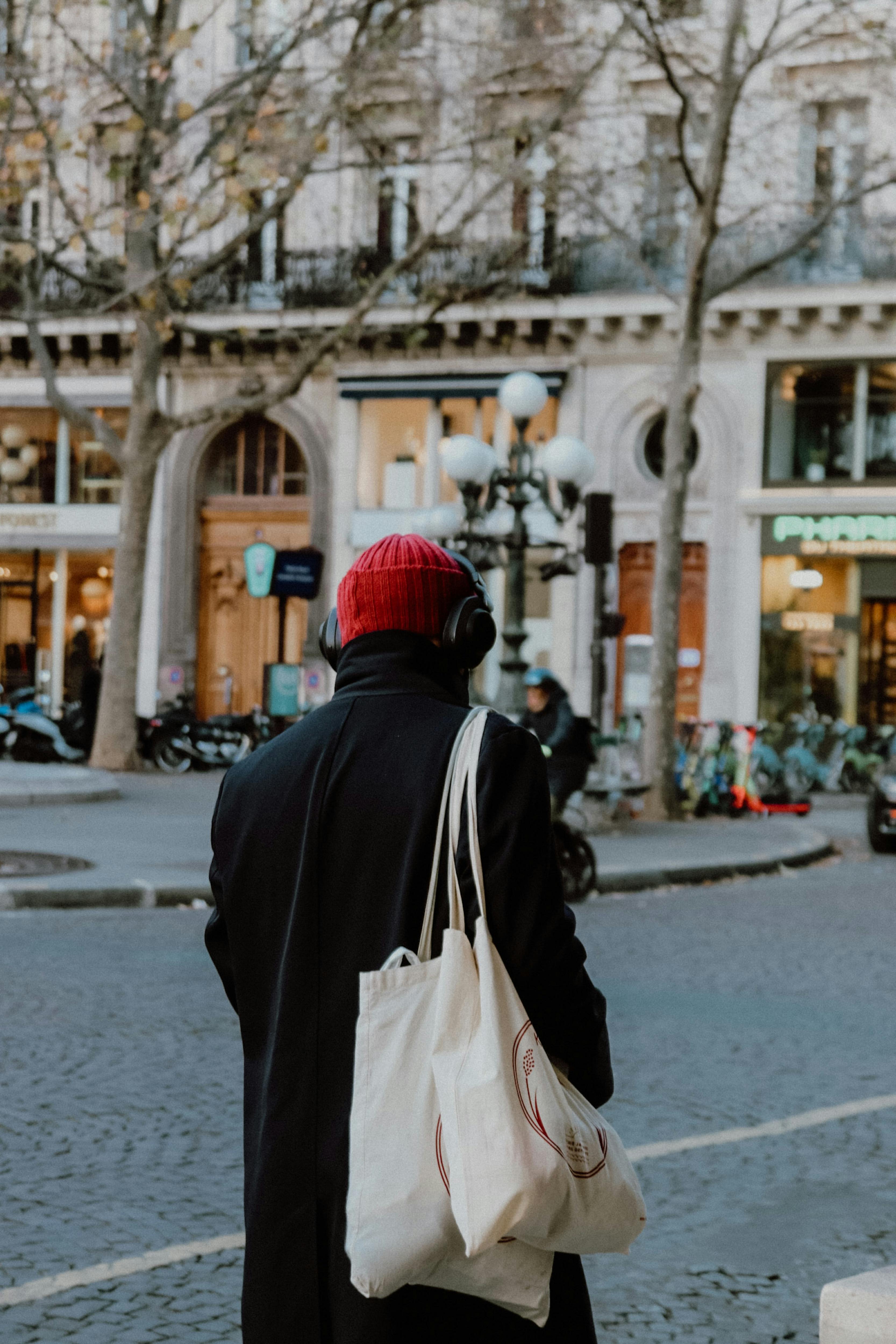 Adult man in a black coat with headphones and tote bag, walking on a cobblestone street.