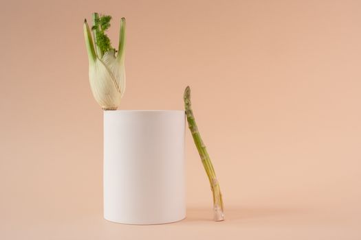 A minimalist still life image featuring asparagus and fennel on a geometric white cylinder against a beige background.