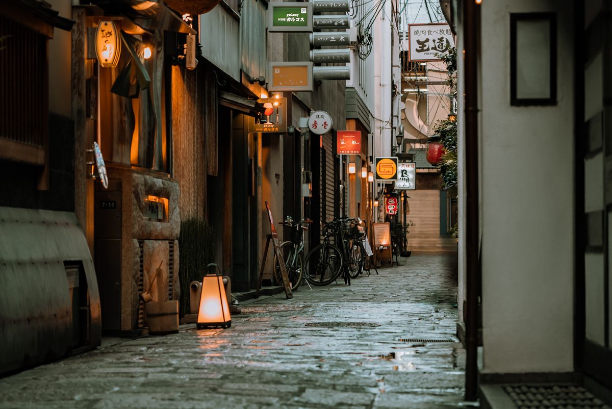 A narrow stone-paved alley in Osaka after dark, red paper lanterns glowing low over shuttered bar fronts, a row of parked bicycles leaning against the wall, the street empty but for lantern light.