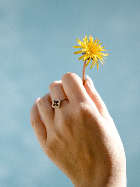Close-up of a hand with a ring delicately holding a yellow flower against a vibrant blue background.