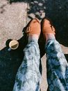 From above of crop anonymous person in stylish trousers and leather loafers sitting on asphalt ground near cup of aromatic coffee