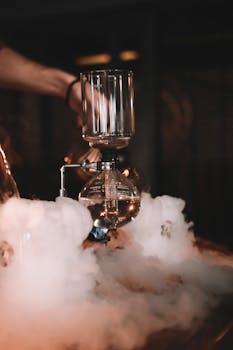 Crop anonymous male barista standing at counter with vacuum coffeemaker and thick vapor in dark cafe