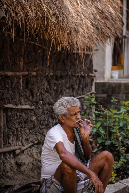 Free An elderly man with gray hair smokes a cigarette while sitting outdoors in a rural setting. He wears a white t-shirt. Stock Photo