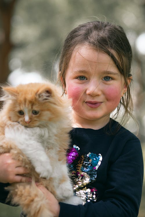 Brown haired little girl with red kitten in hands looking at camera in blurred forest