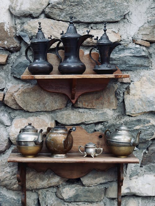 Shiny old fashioned metallic kettles placed on wooden shelves on shabby stone wall on street in daytime