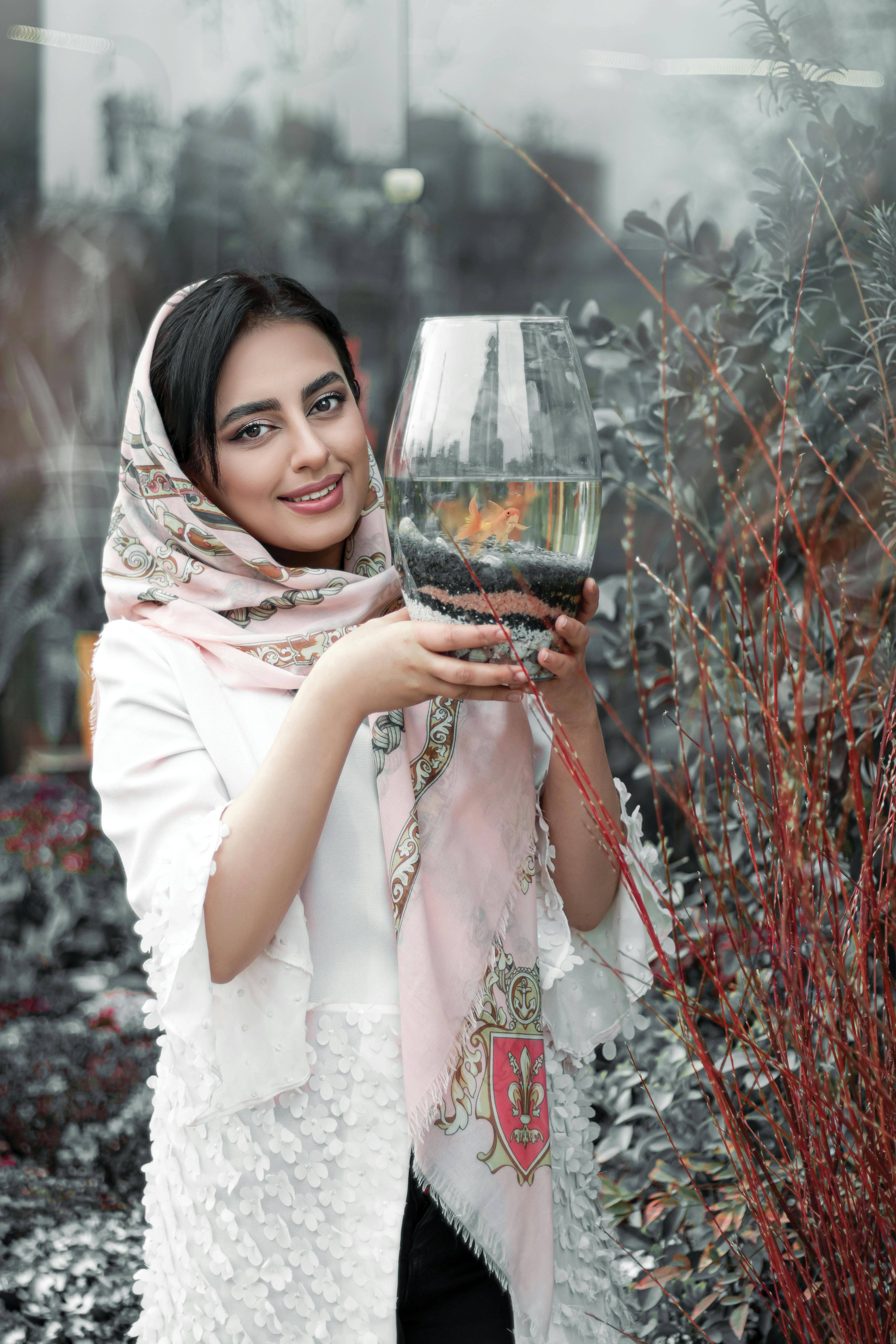 A woman in a white dress and hijab holds a decorative fish jar outdoors, surrounded by foliage.