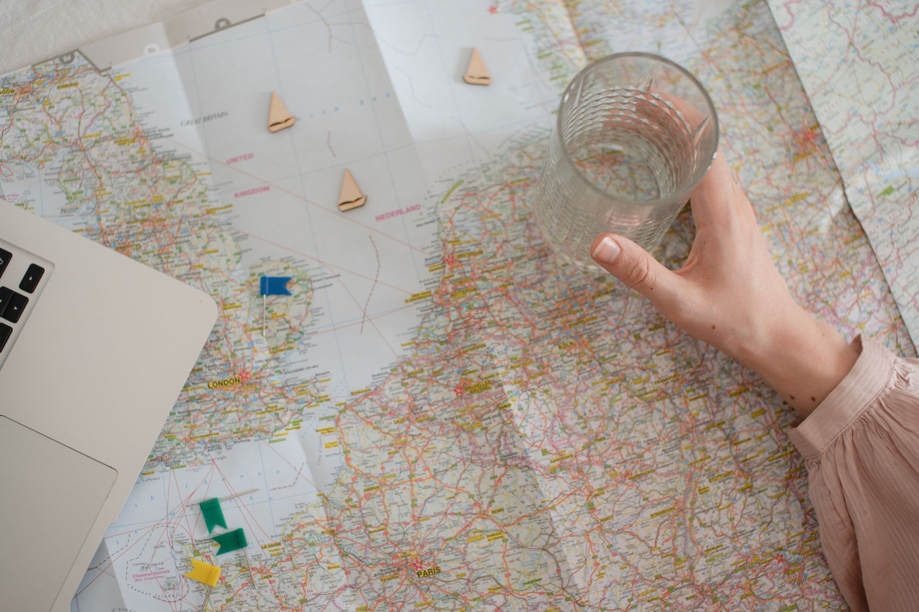 A hand holding a glass over a world map with travel planning items and a laptop.