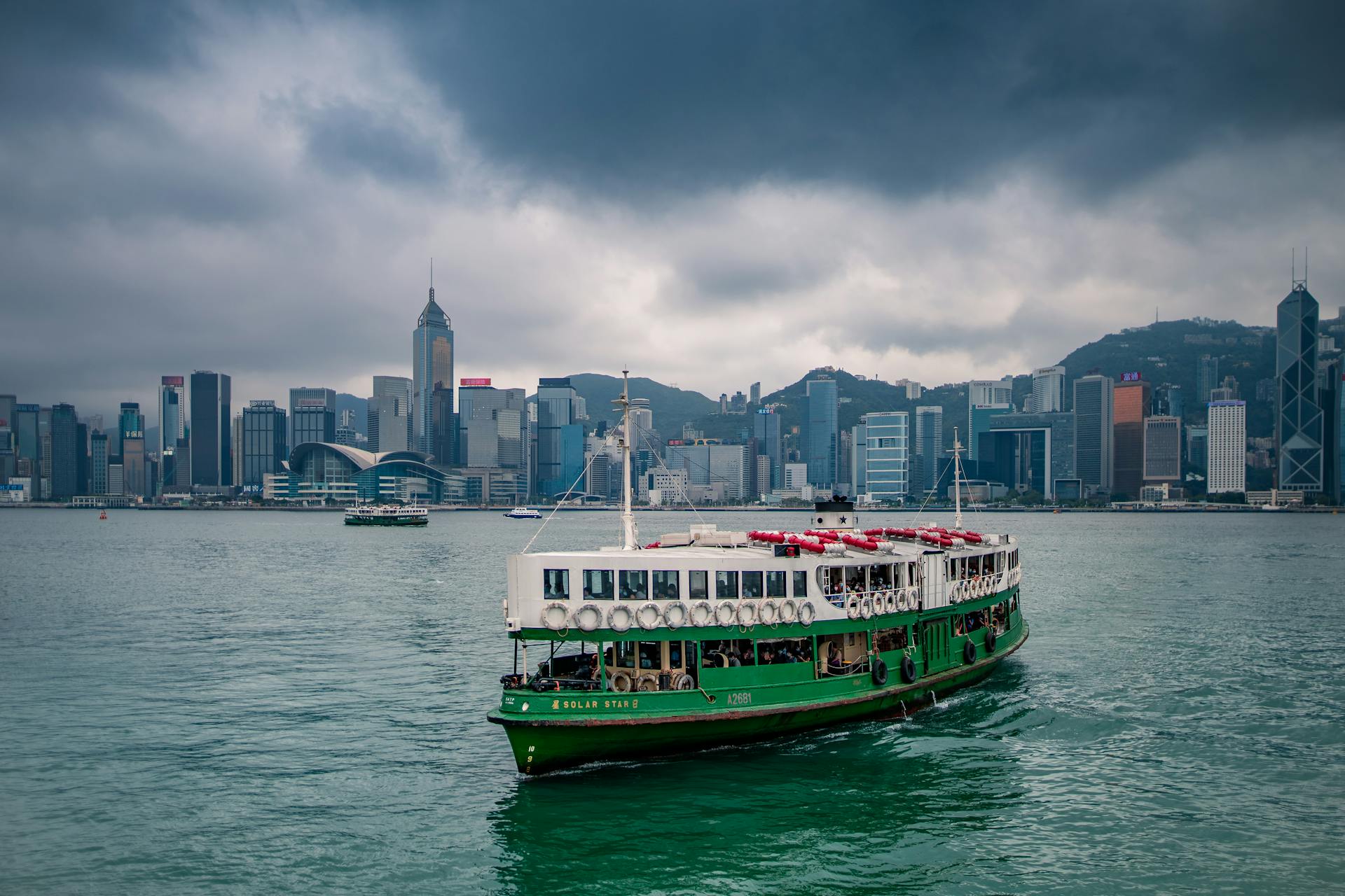 Scenic view of a traditional ferry on Victoria Harbour with Hong Kong skyline under cloudy skies.