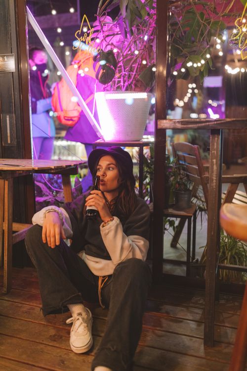 Free A young woman sits casually with a drink, surrounded by colorful neon lights and a lively atmosphere. Stock Photo