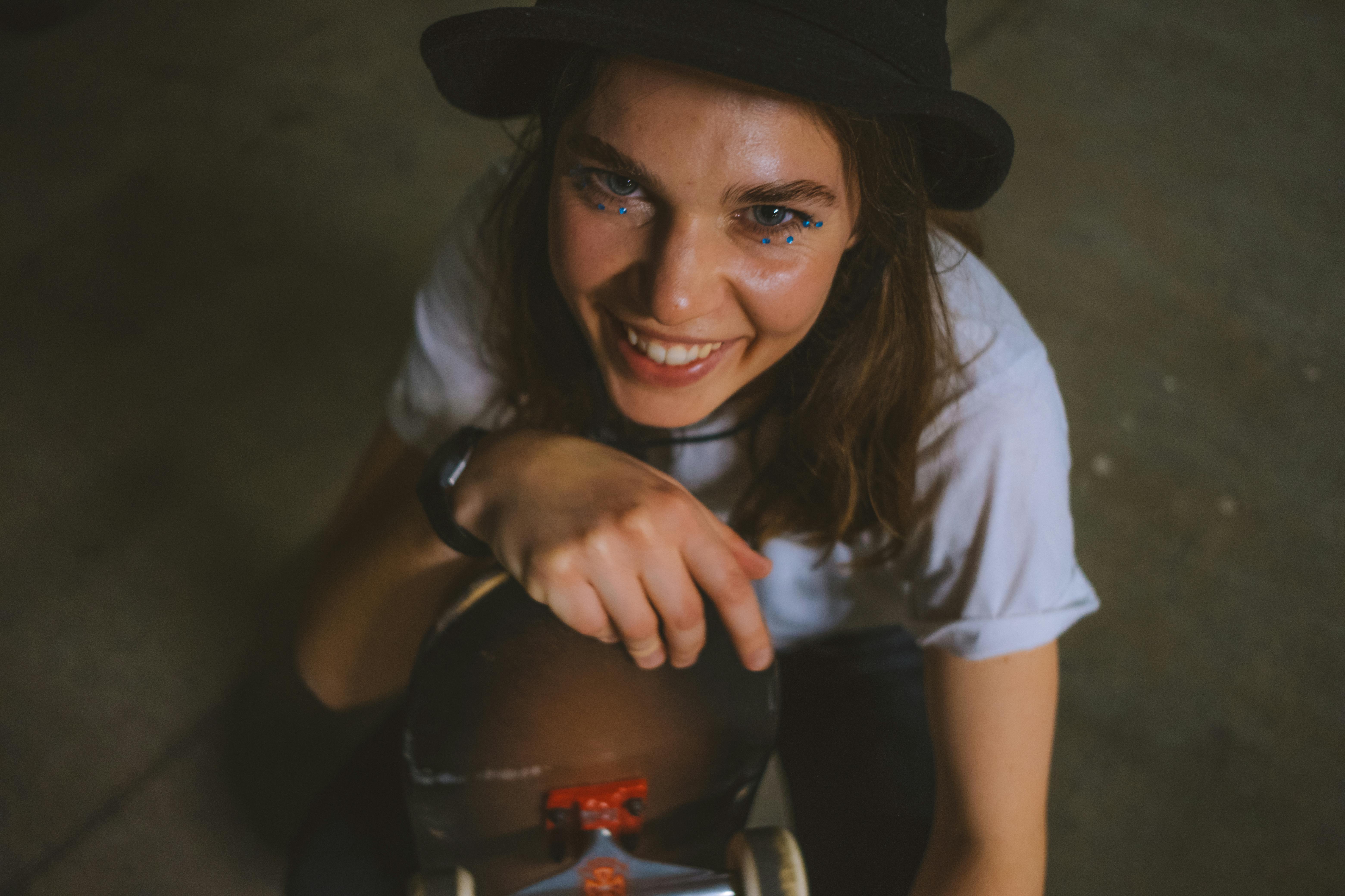 Cheerful female skateboarder sitting indoors holding her skateboard, wearing casual streetwear and a hat.