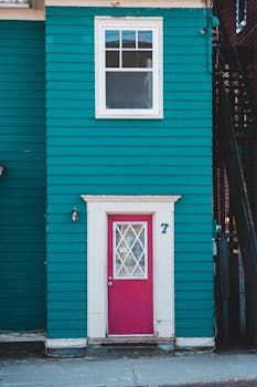 A vibrant teal house featuring a striking pink door and a large white-framed window above.