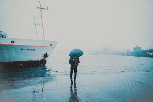 A person in Istanbul stands by a dock in the rain, holding an umbrella near a ship with the city skyline in the background.