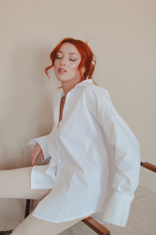 Free Red haired female with closed eyes in white blouse and pants sitting near blank wall Stock Photo