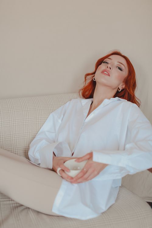 Free Woman with red hair in a white shirt relaxedly holding a ceramic cup indoors. Stock Photo