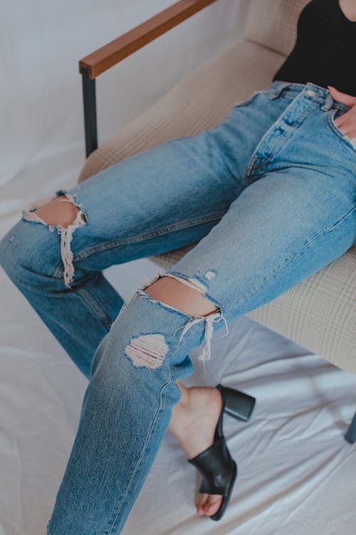 From above of anonymous woman in stylish ripped jeans and leather mule shoes sitting on chair placed on sheet