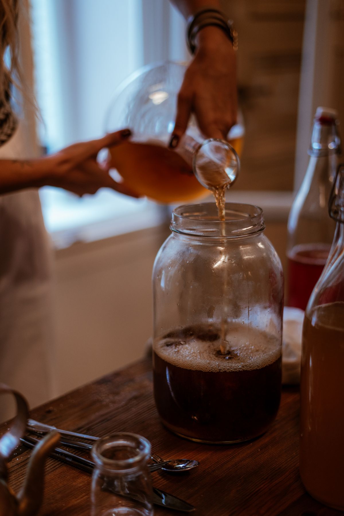 A person pouring home-brewed kombucha from a large glass fermentation jar on a wooden kitchen table
