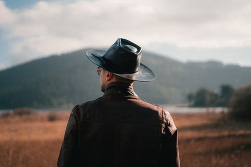 Free A man wearing a cowboy hat stands in a field, gazing at distant hills under a clear sky. Stock Photo