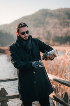 A man in a coat pours whiskey into a glass with scenic outdoor backdrop.