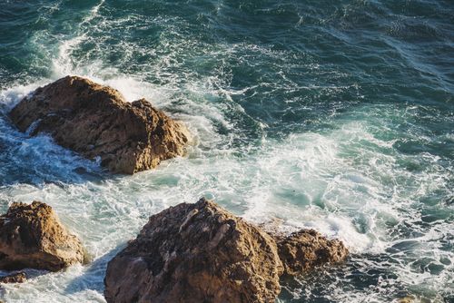 Free An aerial view capturing the dynamic waves crashing against rugged coastal rocks in a sunlit seascape. Stock Photo