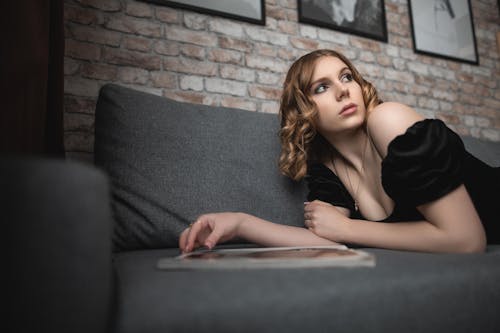 Free From below of thoughtful young woman with hairstyle in stylish clothes lying on comfortable sofa and looking away against brick wall with paints in room Stock Photo