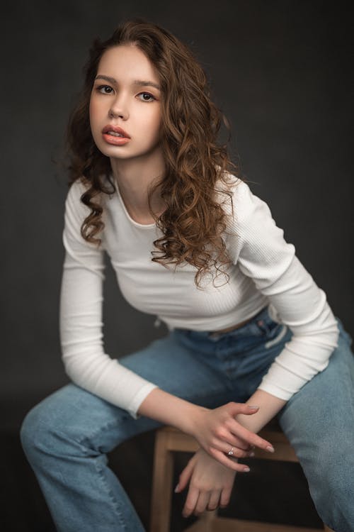 Free Pensive young female model with curly brown hair in trendy clothes sitting on chair and looking at camera against dark background in studio Stock Photo
