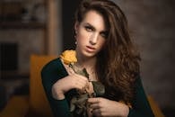 Young female with long hair in green clothes sitting in yellow armchair with yellow rose and looking at camera