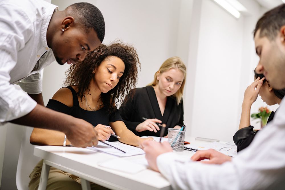 Quatre personnes travaillent ensemble autour d'une table blanche dans un bureau lumineux, examinant des documents et prenant des notes lors d'une réunion collaborative.