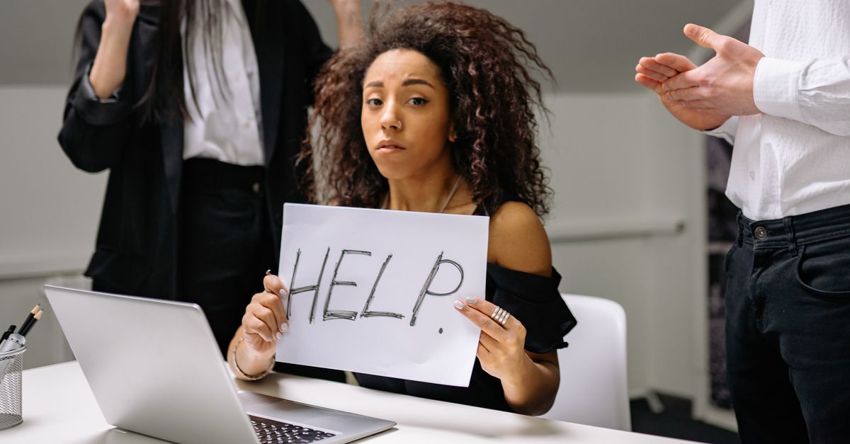 An unhappy businesswoman showing a sign reading 'HELP' in an office setting.